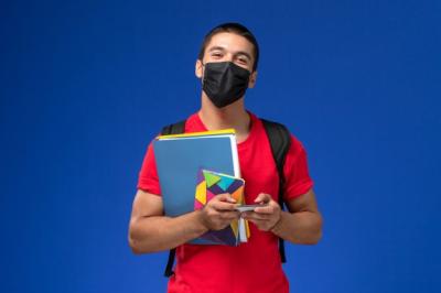 estudiante-masculino-vista-frontal-camiseta-roja-mochila-mascara-sosteniendo-archivos-usando-su-telefono-fondo-azul_140725-40825